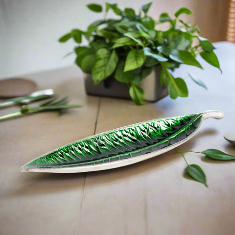 An aluminum metal serving tray with a mango leaf shape design, displayed on a table with a plant in the background.